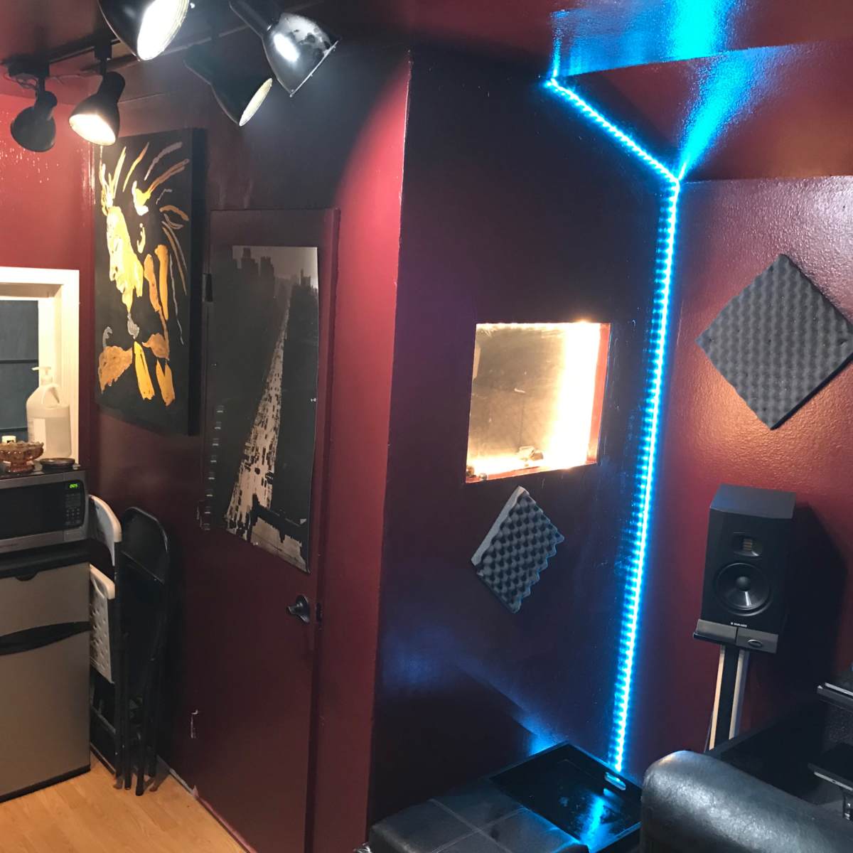 A room with maroon walls, featuring a small kitchen area with a fridge, a wall art piece, and a corner highlighted by blue LED lights.