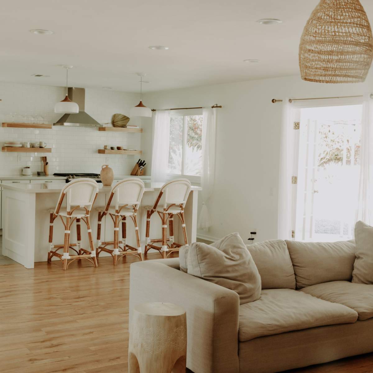 A modern living room features a light-colored sofa and a kitchen with bar stools, hardwood flooring, and ample natural light.