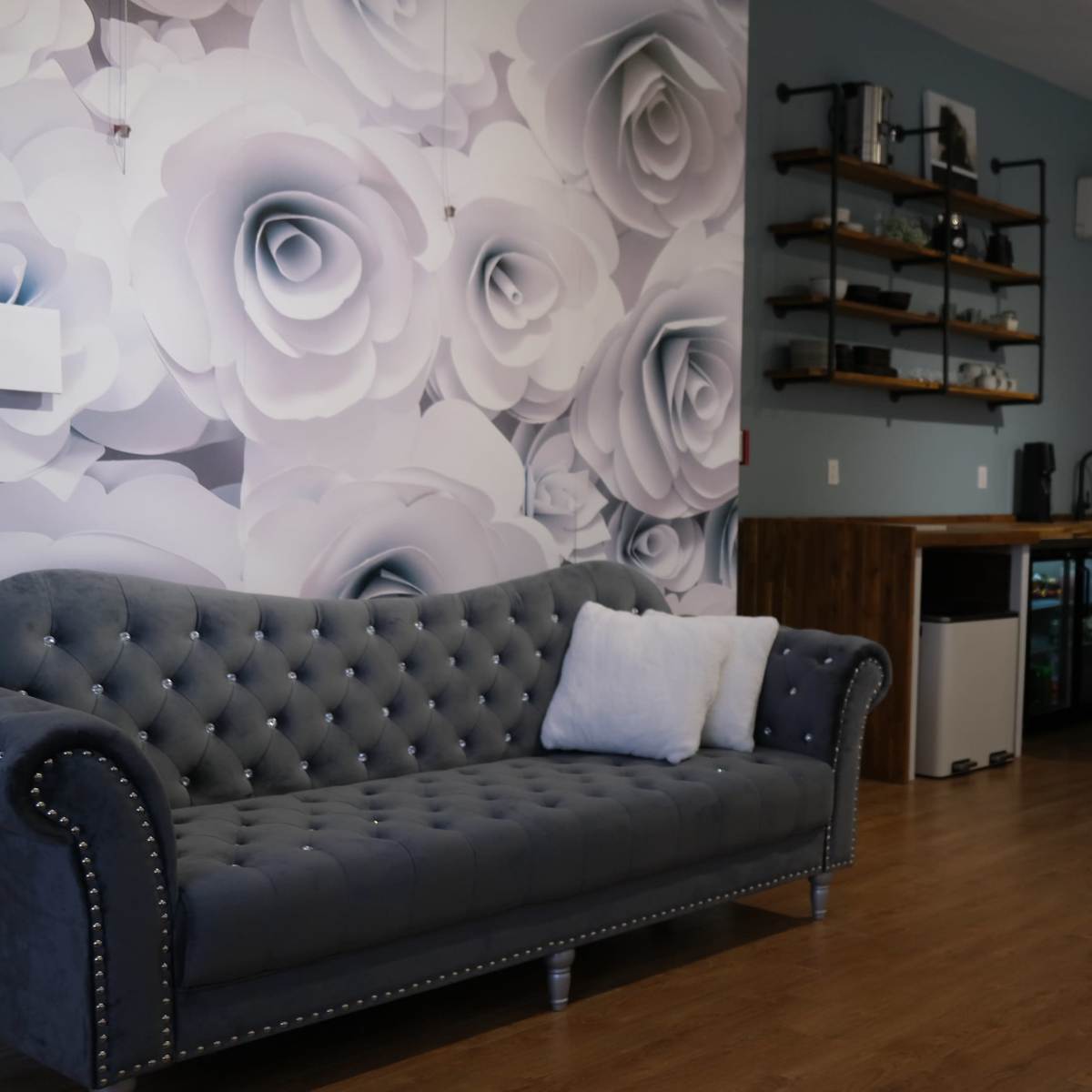 A modern interior featuring a gray tufted sofa, a floral wall mural, a wooden bar area with stools, and a small kitchen setup in a spacious room with hardwood flooring.