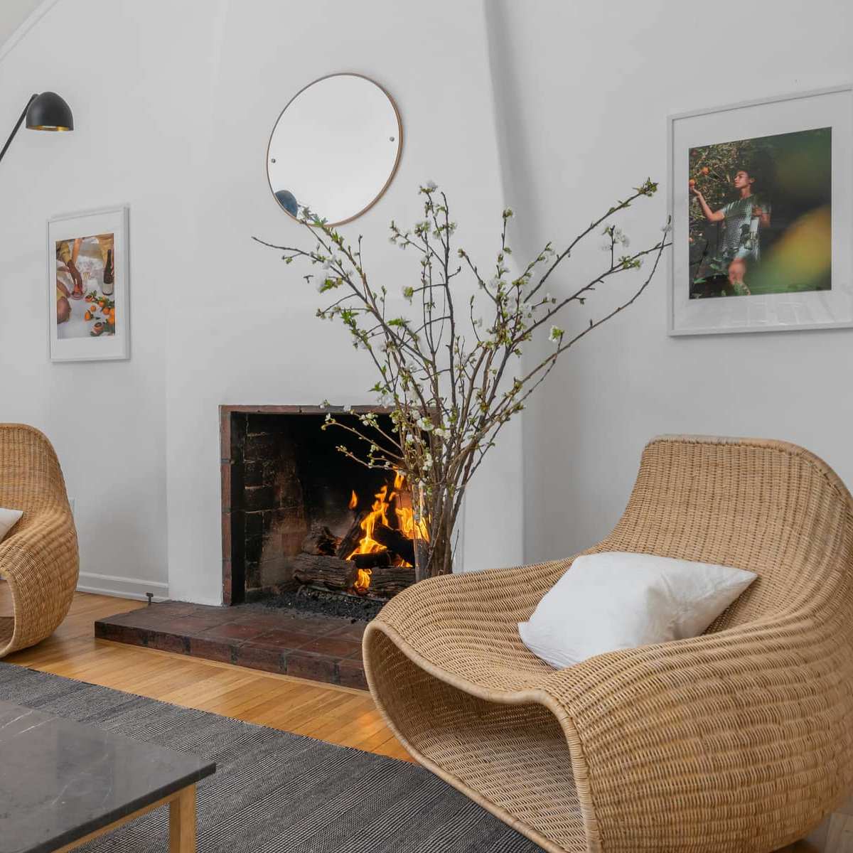 A minimalist living space with two woven chairs, a coffee table, a fireplace with a fire burning, and framed artwork on the walls.