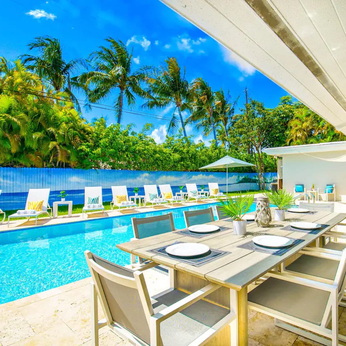 An outdoor dining area with a wooden table set for a meal, overlooking a swimming pool surrounded by lounge chairs and palm trees.