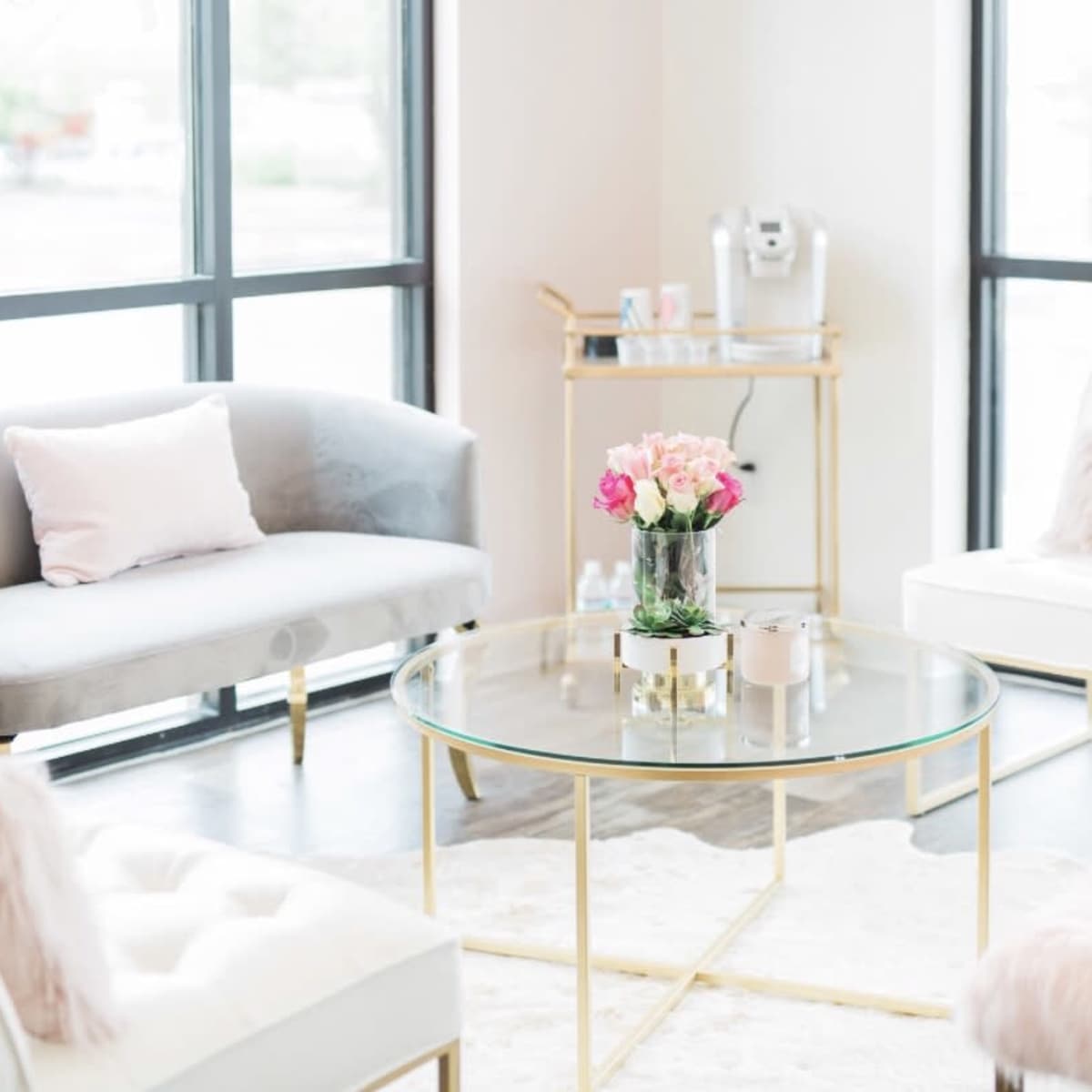 A modern waiting room featuring a glass coffee table surrounded by a gray couch and two white chairs, with a bouquet of flowers on the table.