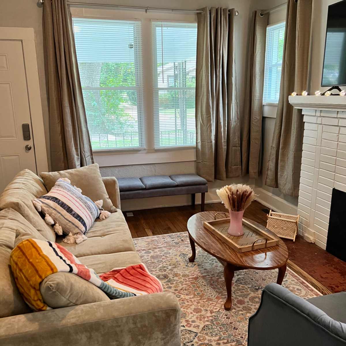A living room with a beige sofa, a blue armchair, a wooden coffee table, and a white brick fireplace, all complemented by large windows draped with beige curtains.