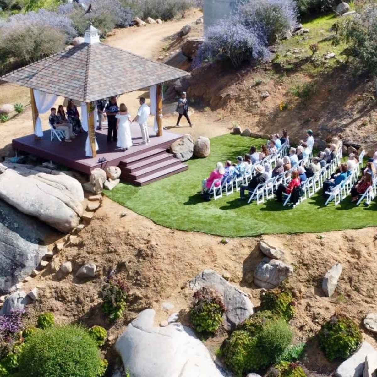 A wedding ceremony takes place under a gazebo on a hillside, surrounded by guests seated in rows on green grass.