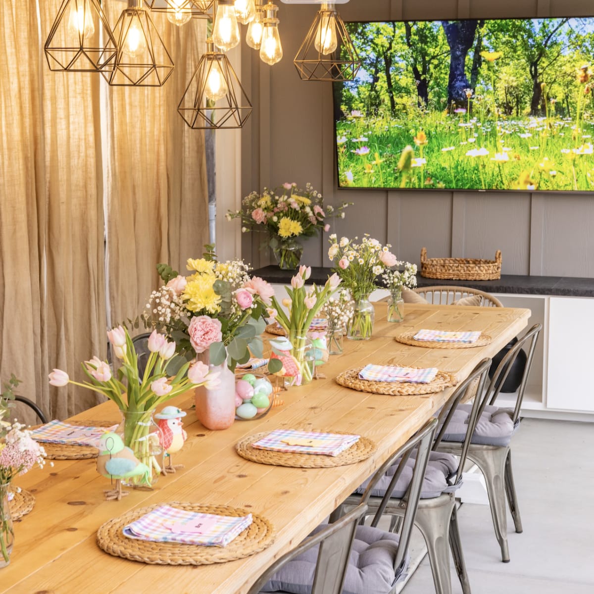 A dining area features a long wooden table set with pastel-colored tableware and surrounded by wire-framed chairs, with a large screen displaying a nature scene in the background.