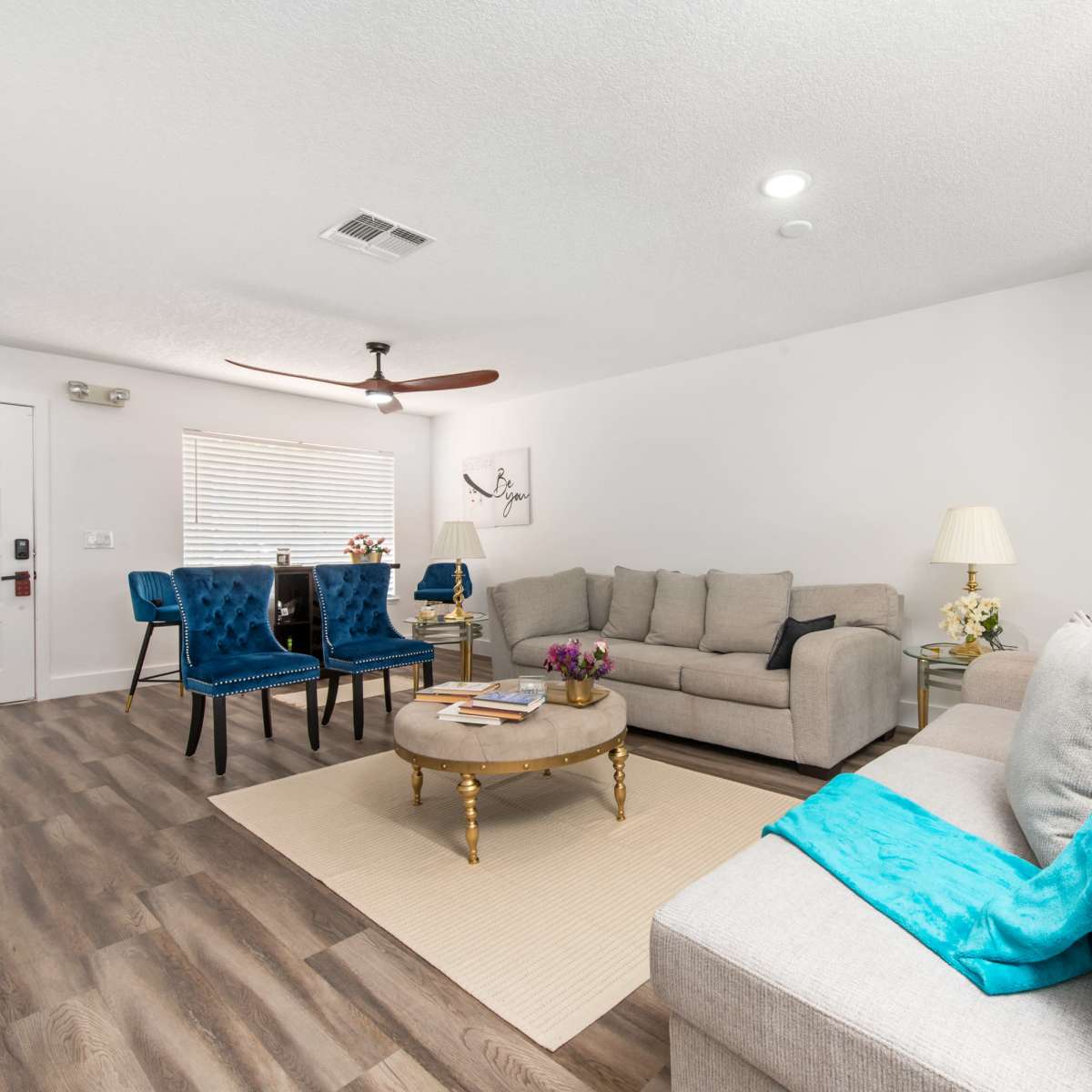 A living room features two sofas, a coffee table, and a dining area with blue chairs, illuminated by natural light.