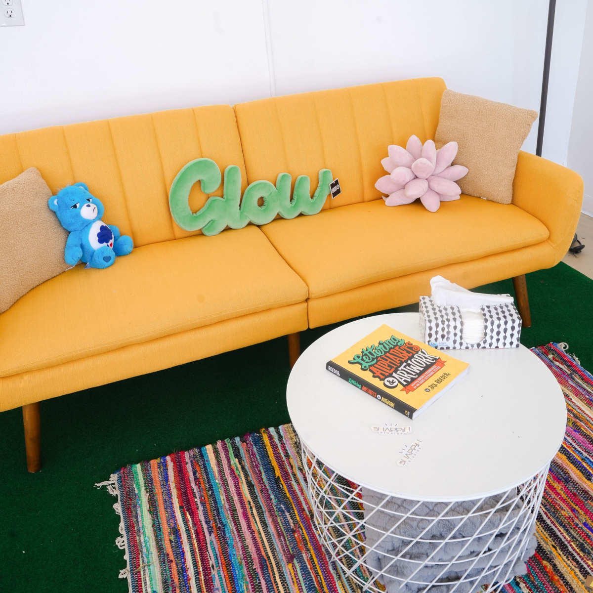 A bright yellow couch with decorative pillows and a green "Glow" sign sits beside a white round coffee table on a colorful woven rug.