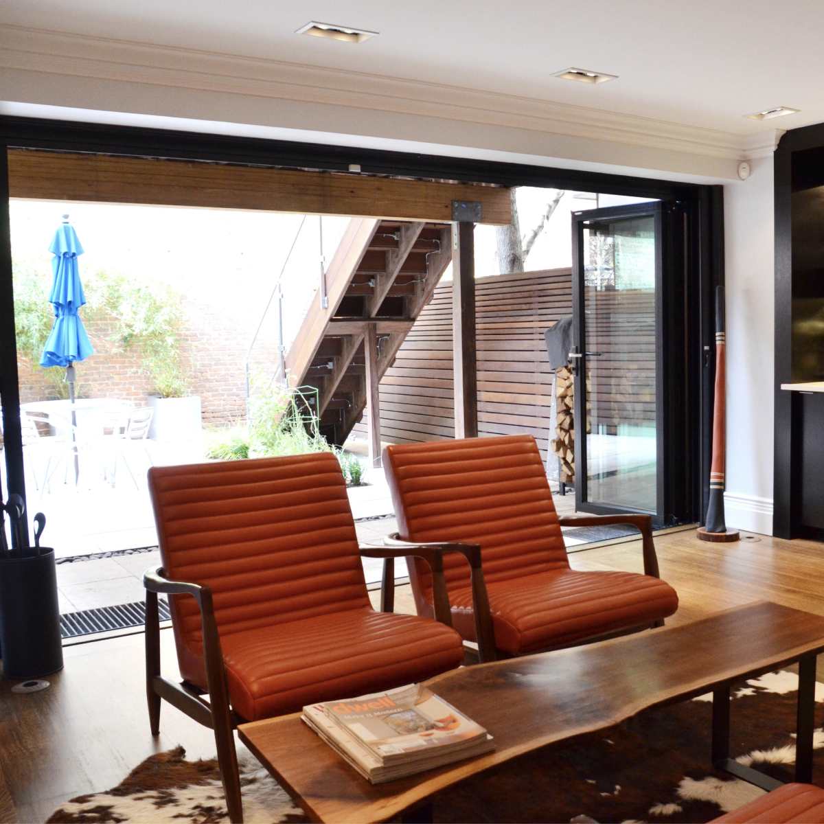 A modern living space with two leather chairs, a wooden coffee table, and a bar with glassware, featuring large glass doors that open to an outdoor patio area.