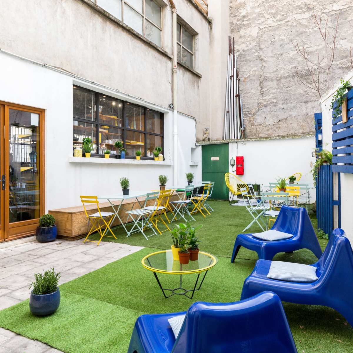 An indoor-outdoor space with a green lawn, blue chairs, yellow tables, and potted plants, bordered by a beige wall and a wooden door.