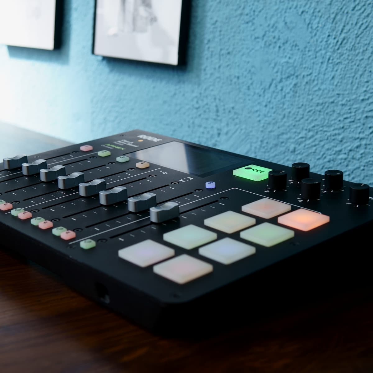 A music production device with various buttons and sliders, placed on a wooden surface against a blue wall with framed artwork above.