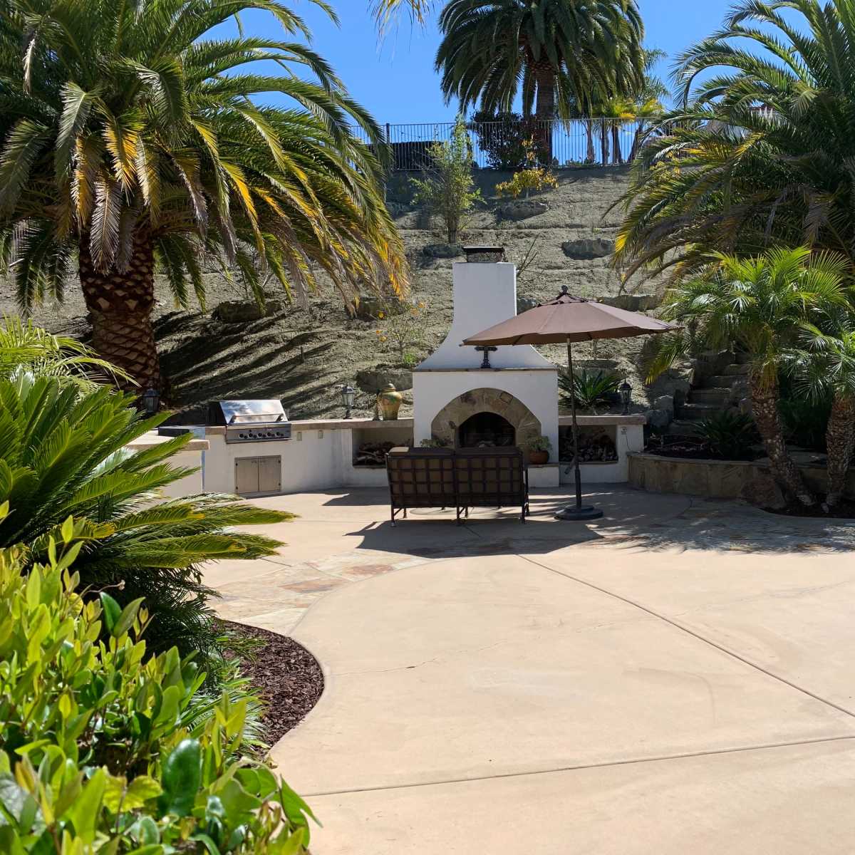 A landscaped patio area with palm trees, an outdoor fireplace, a shaded seating arrangement, and a swimming pool.