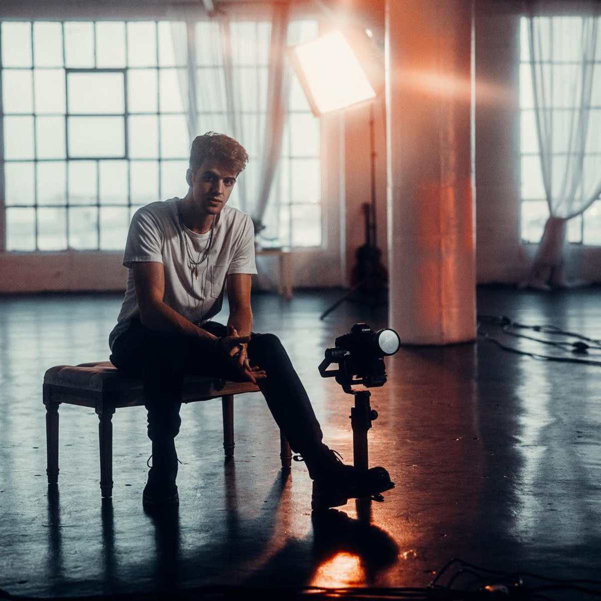 A young man sits on a stool in a well-lit studio with large windows and a camera positioned on a tripod nearby.