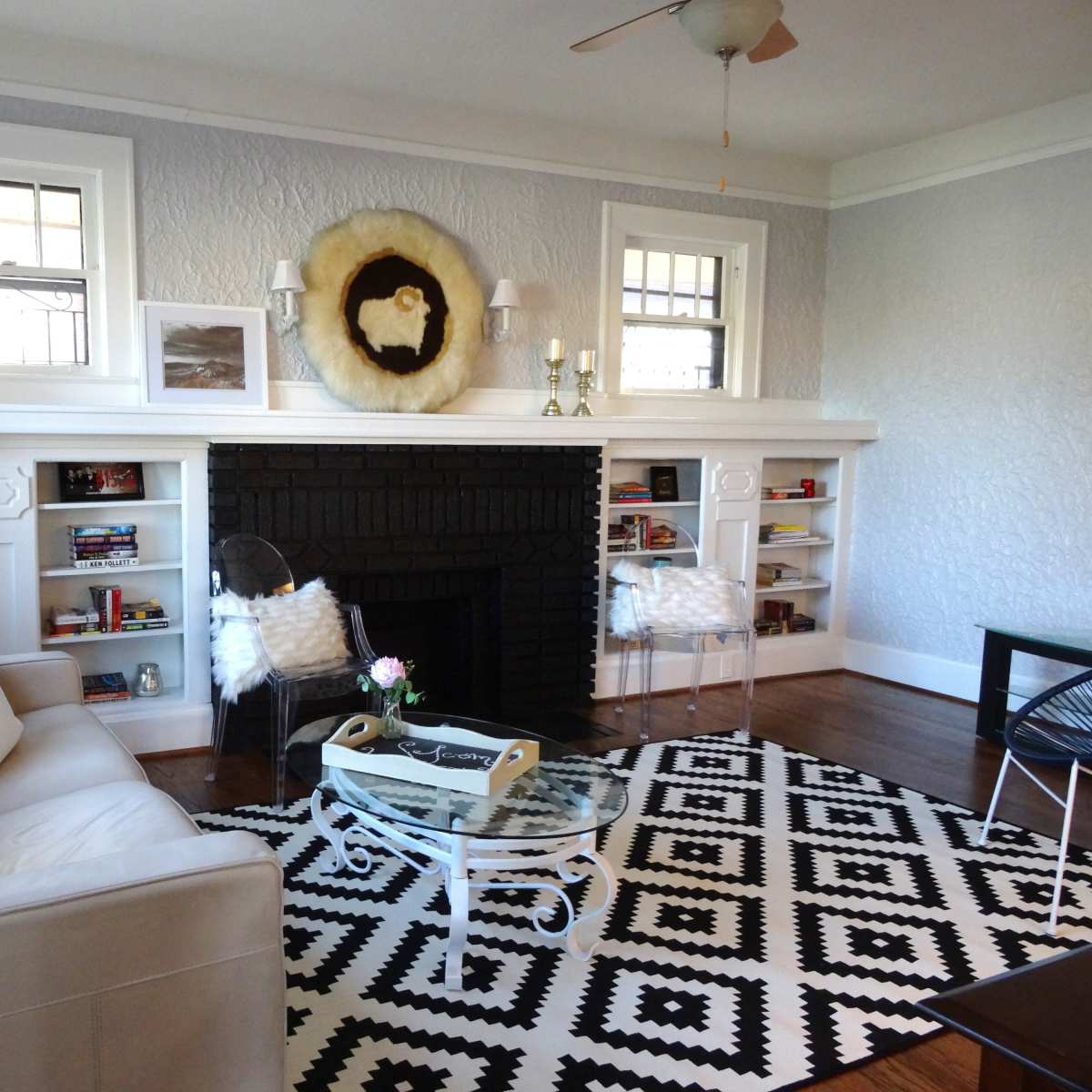 A living room features a black fireplace surrounded by built-in shelves, a glass coffee table on a patterned rug, and a white sofa accompanied by two fluffy chairs.
