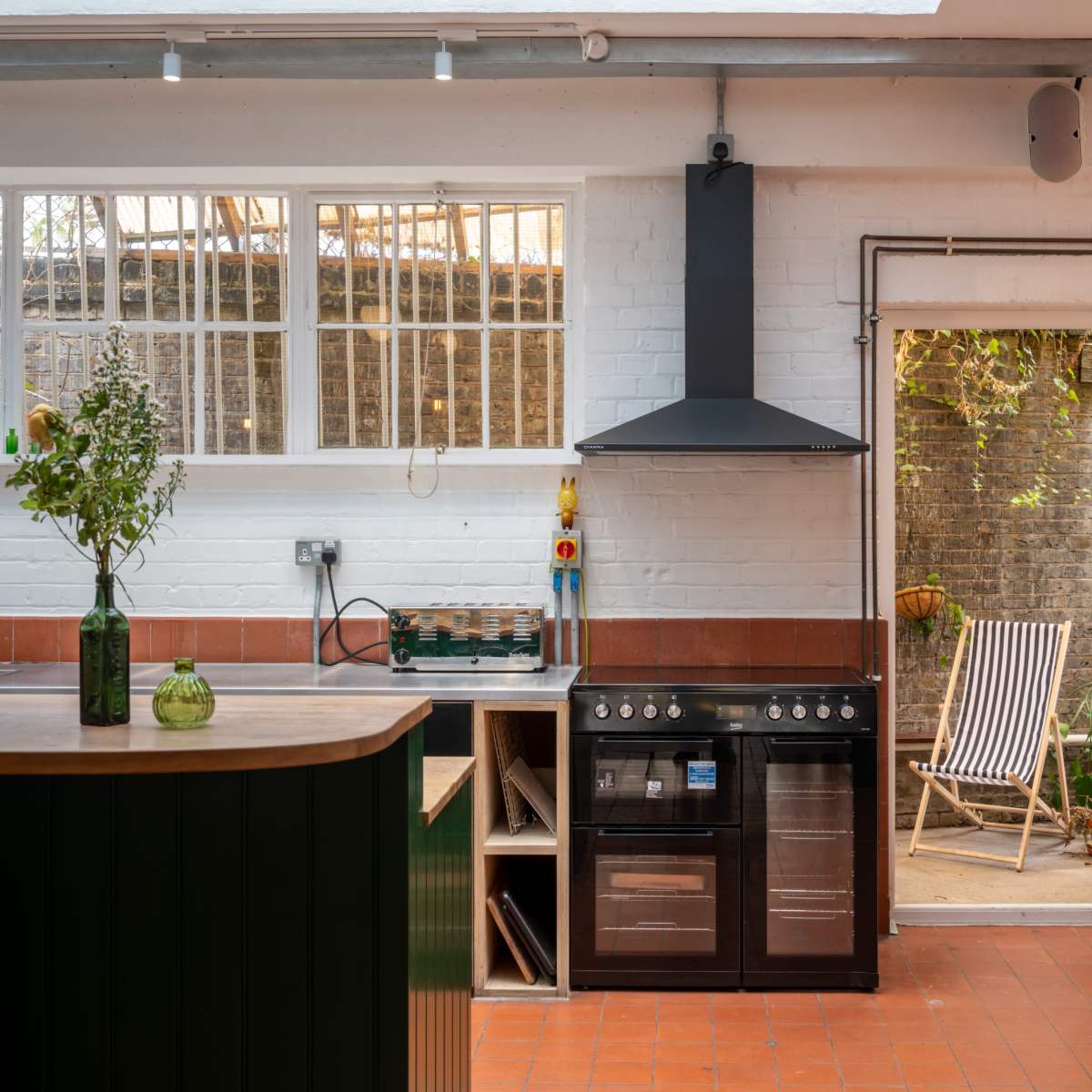 A modern kitchen with a black stove, a wooden counter, and a door leading to a garden with a striped lounge chair.