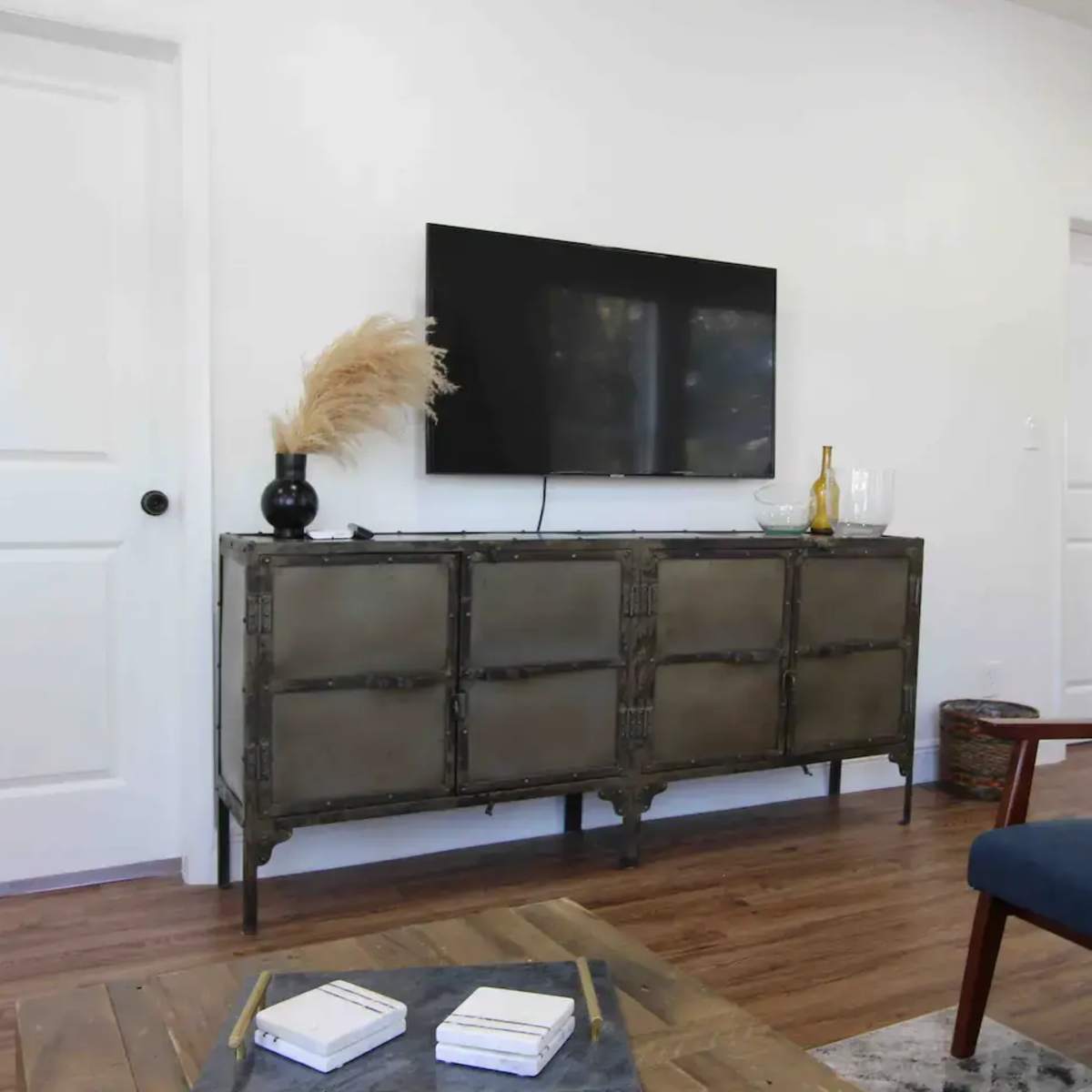 A living room with a wall-mounted TV above a metallic television stand, accompanied by a blue armchair and a wooden coffee table.