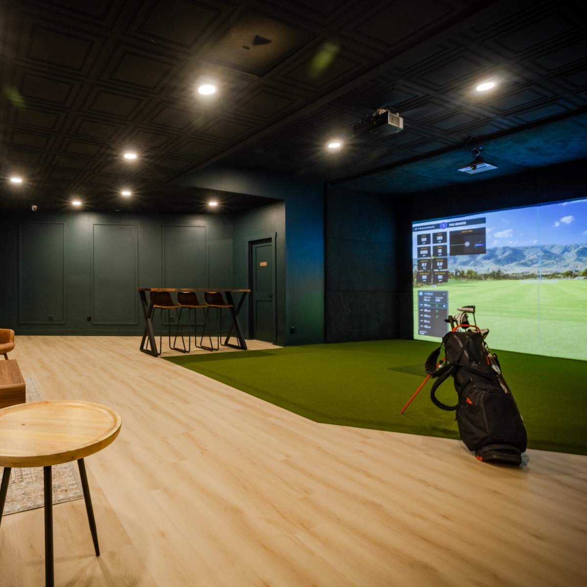 A modern indoor golf simulation room featuring a putting area, a lounge with comfortable seating, and a large projector screen displaying a golf course.