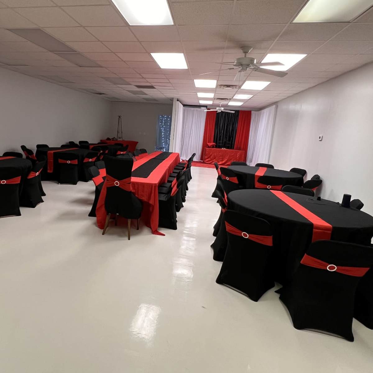 A decorated event space with several round tables covered in black tablecloths featuring red accents, arranged in a spacious, brightly lit room.