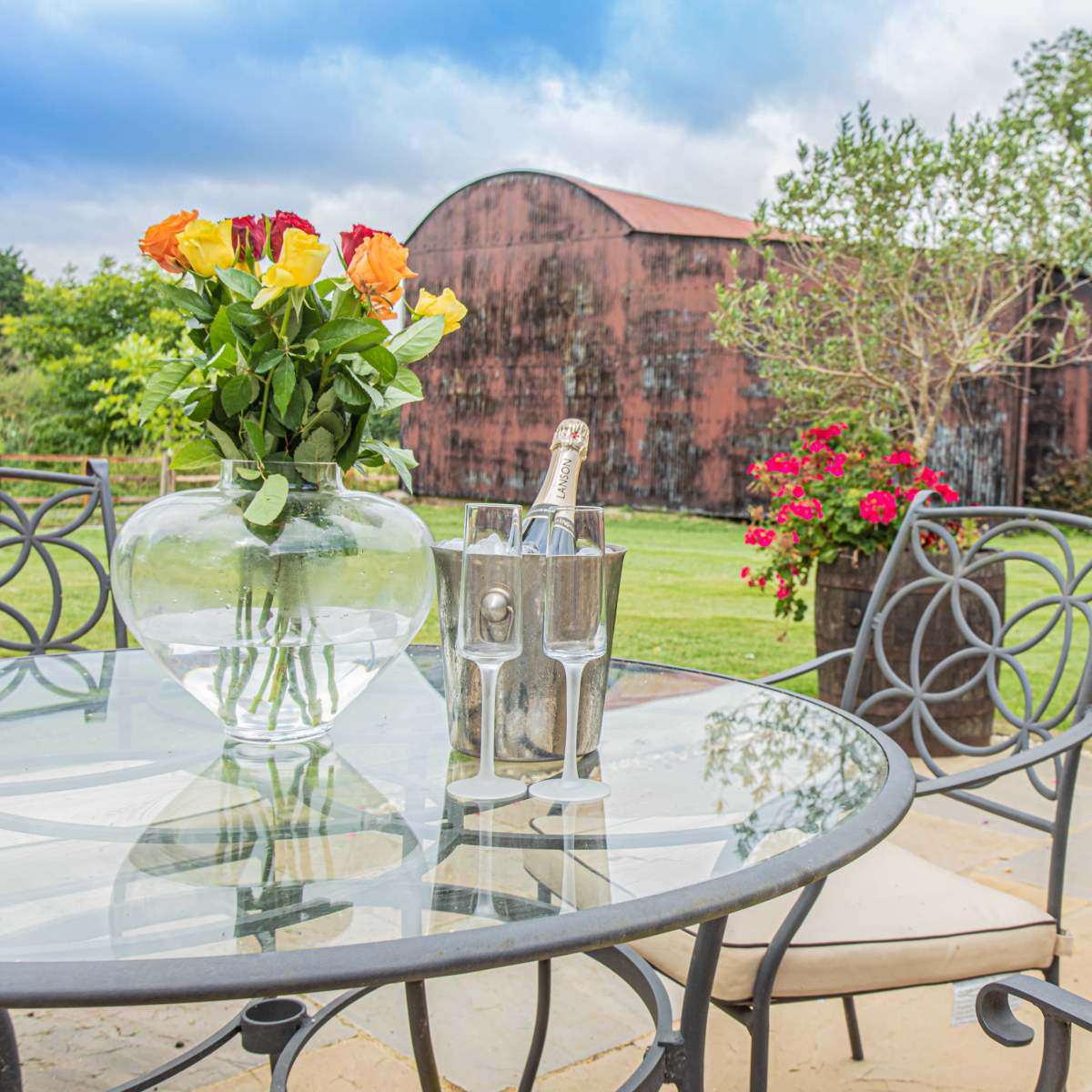 A glass table with a bouquet of roses and a bottle of champagne in an ice bucket is set on a patio overlooking a grassy area and a barn in the background.