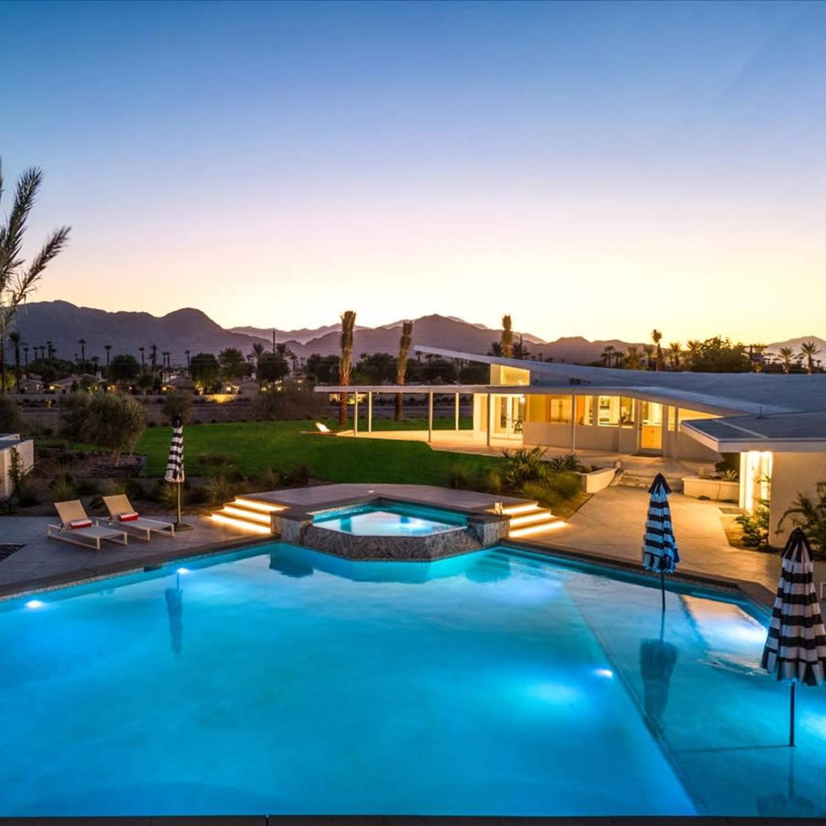 A modern swimming pool with a spa, surrounded by lounge chairs and palm trees, set against a backdrop of mountains at sunset.
