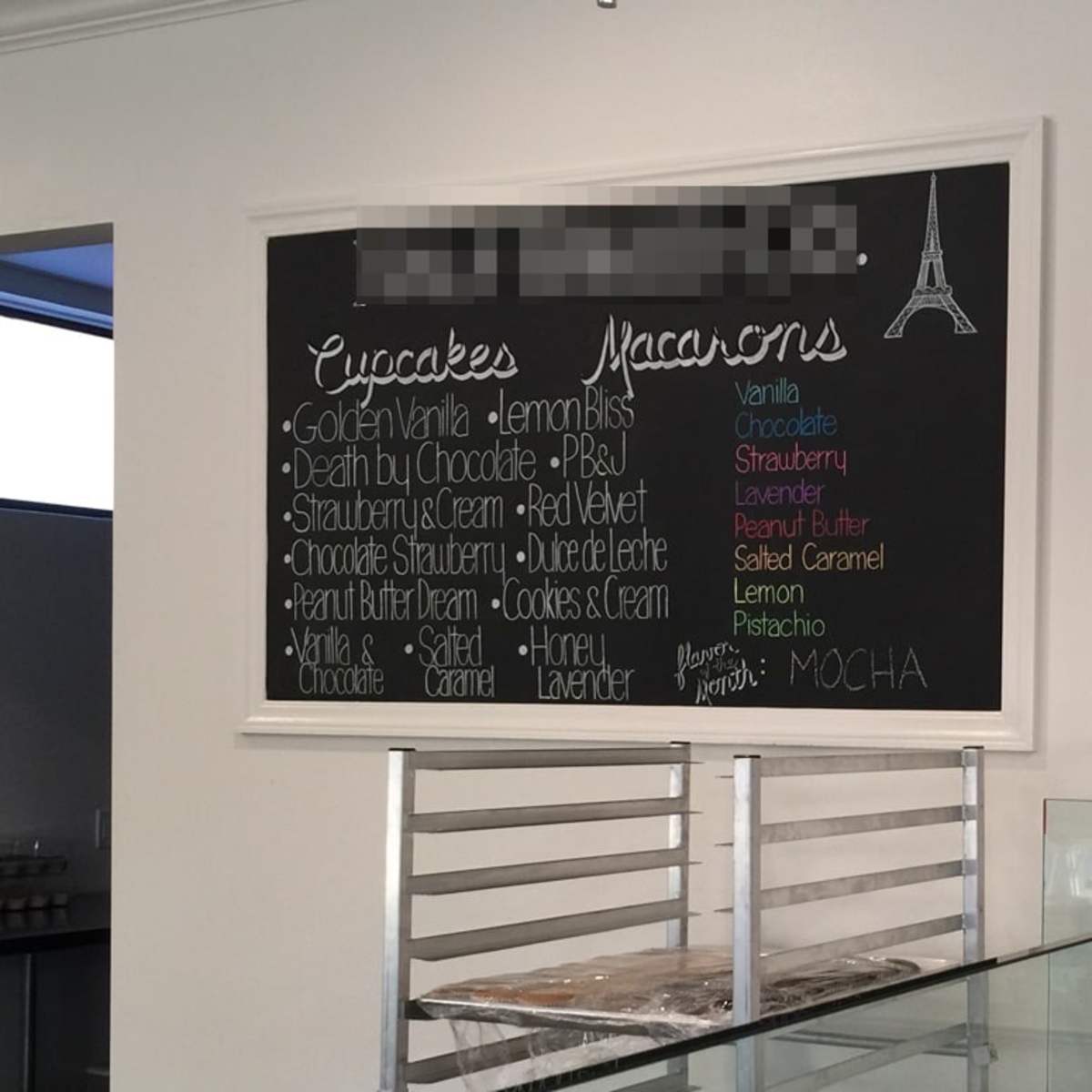 A menu board displays various flavors of cupcakes and macarons in a bakery setting.