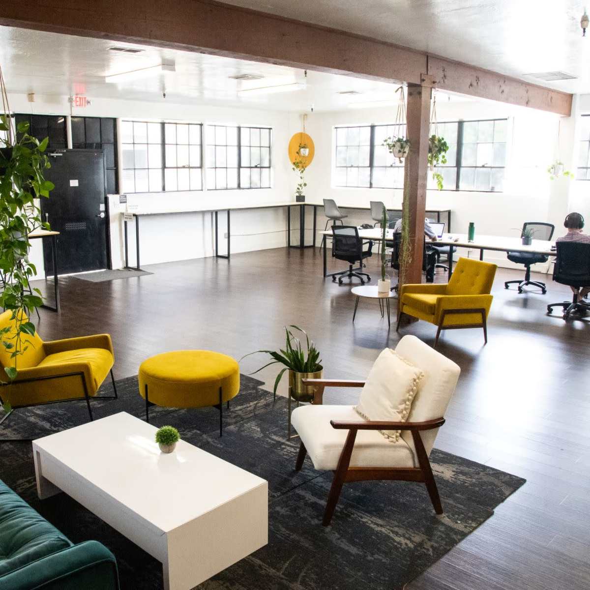 A modern, open office space with a seating area featuring yellow and white chairs, a dark green sofa, and several plants, alongside workstations with desks and computers.