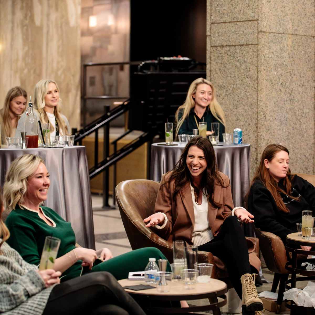 A group of women sits in stylish chairs around small tables in a modern indoor space, engaging in conversation and enjoying drinks.