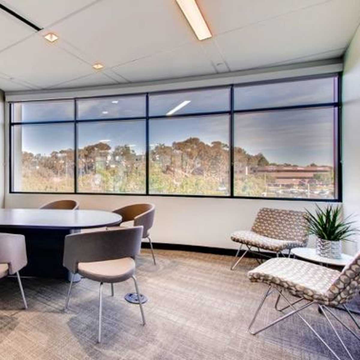 A modern meeting room with a round table, four chairs, a wall-mounted television, and large windows overlooking a green landscape.