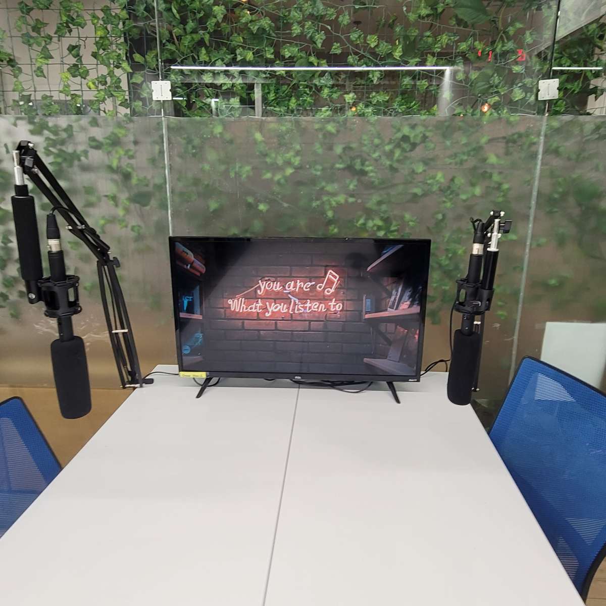 A conference table with two microphones and a television displaying the text "you are what you listen to," set against a backdrop of greenery.
