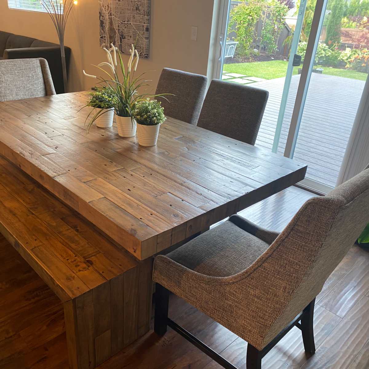 A wooden dining table with a bench and chairs is positioned in a well-lit room near a sliding glass door that opens to a garden.