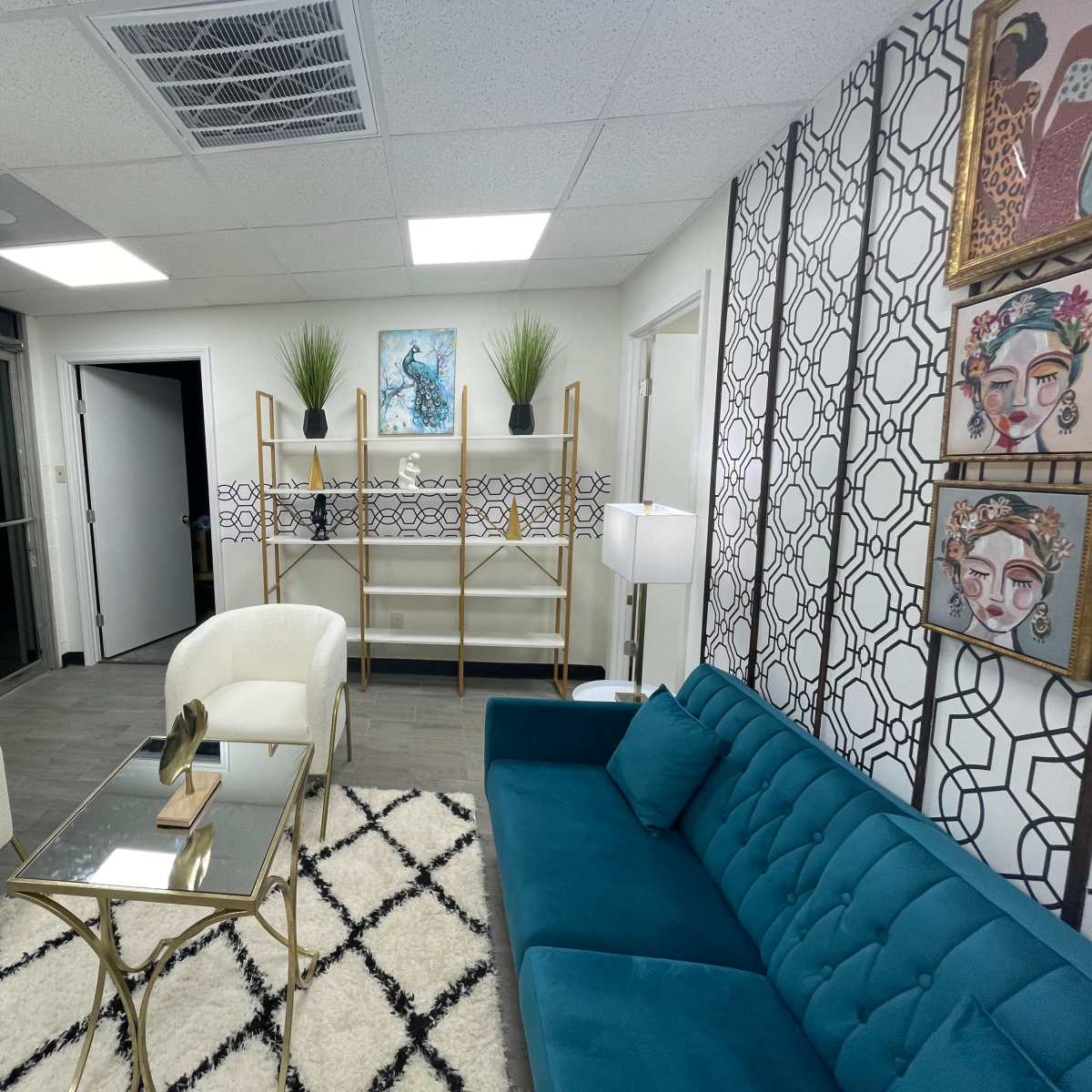 A modern lounge area with a teal sofa, two white armchairs, a glass coffee table, and decorative wall art, featuring a shelving unit and plants.
