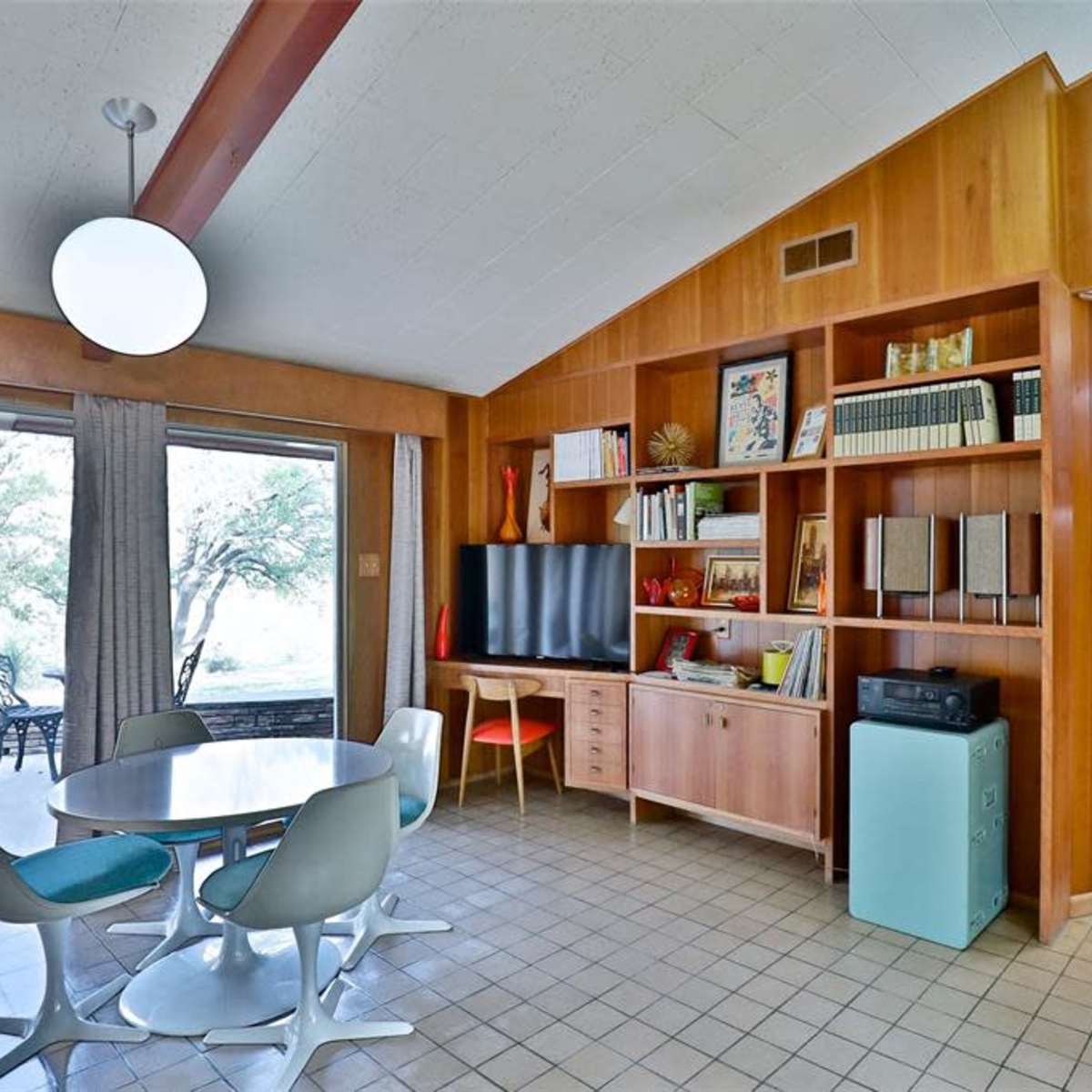 A mid-century modern interior features a dining area with a round table and chairs, a wooden bookshelf with various items, and a television against a paneled wall, leading to a view of the outdoors through large sliding glass doors.