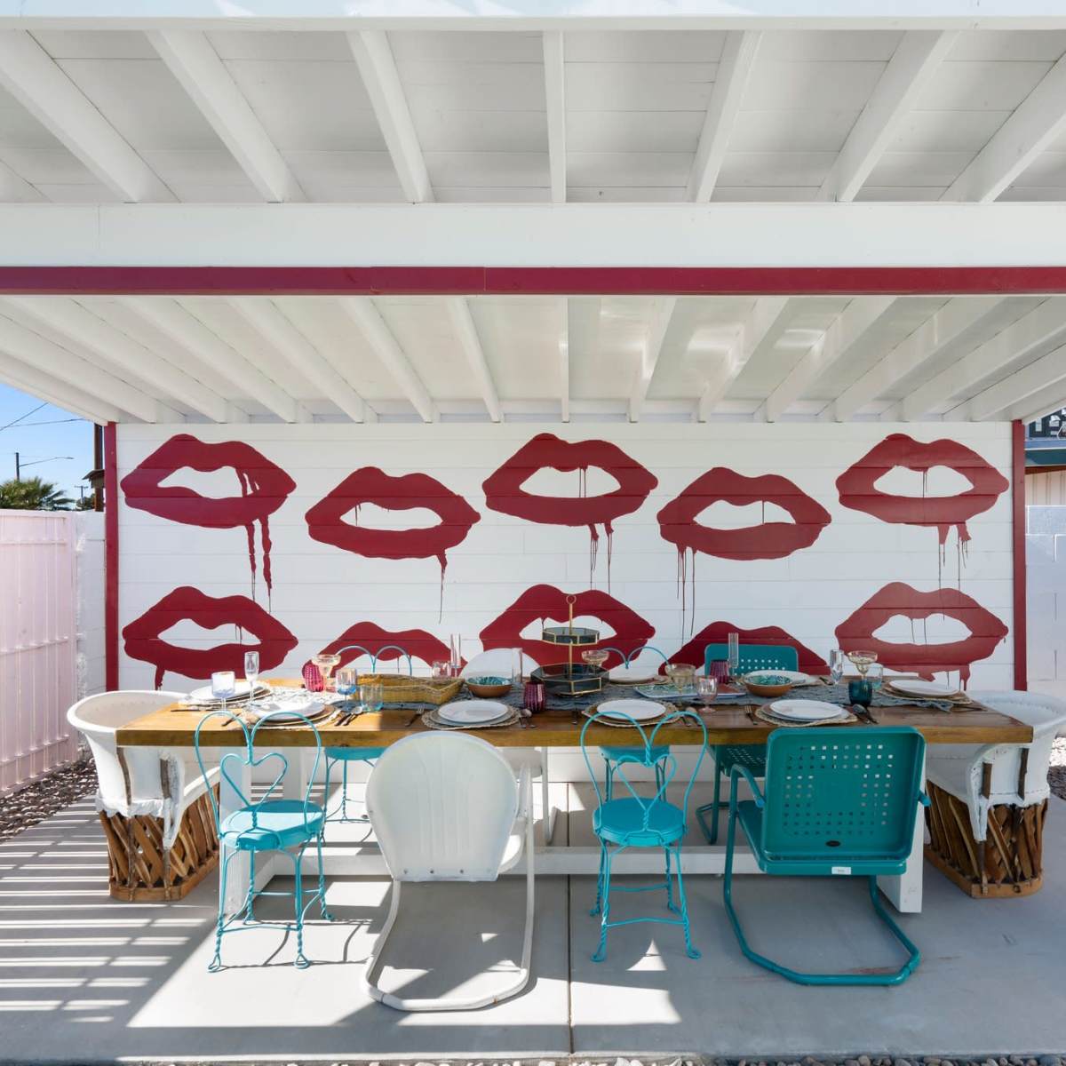 An outdoor dining area under a covered patio with a large table set for a meal and decorated with red lip prints on the walls.