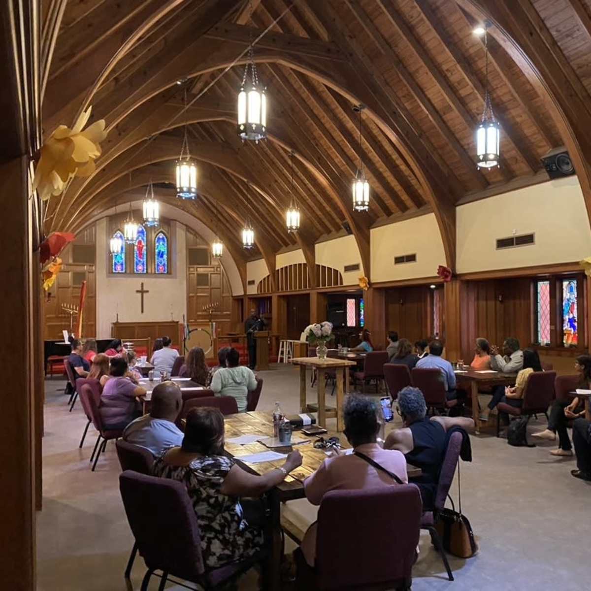 A group of people is seated at tables in a church with wooden beams, stained glass windows, and decorative elements.