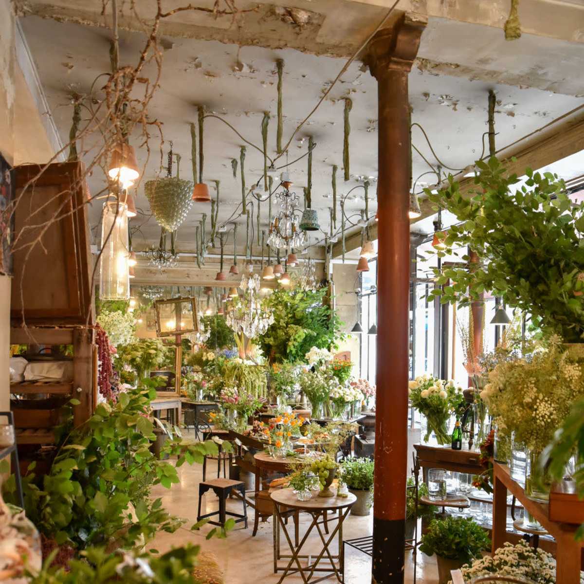 The interior of a floral shop features various plants and flowers arranged on tables, with exposed beams and an industrial-like ceiling.