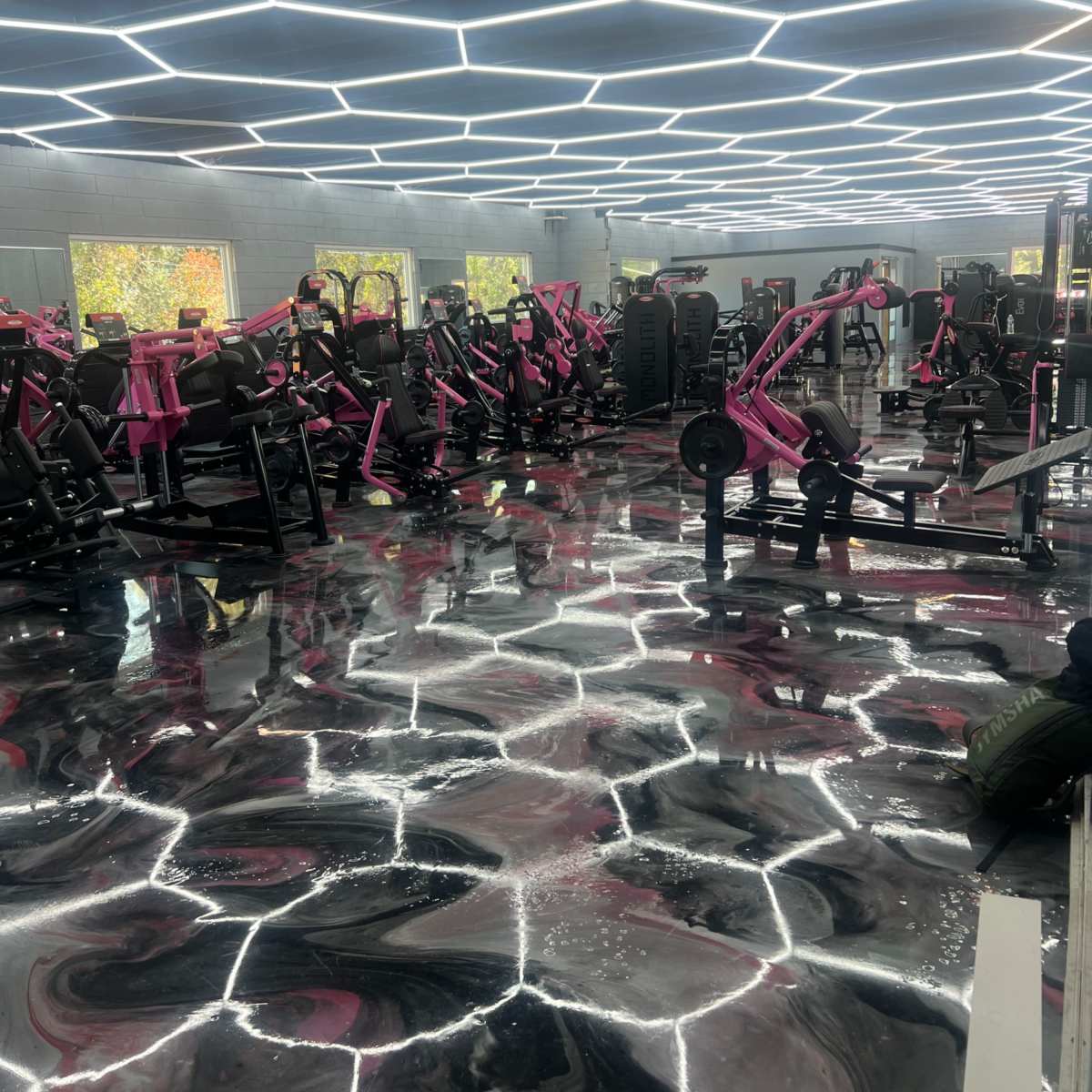 A modern gym interior featuring an array of fitness equipment in shades of pink, complemented by a glossy black and white floor and bright geometric lighting.