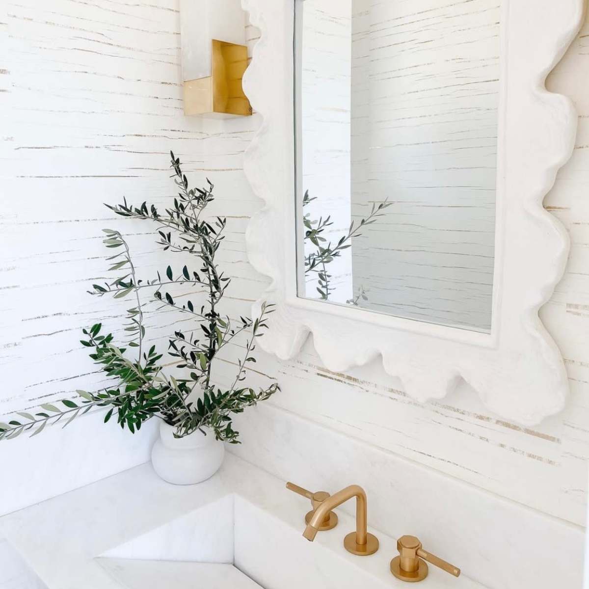 The bathroom features a white textured wall, a scalloped mirror, a gold sconce, and a sink with gold faucets alongside a potted plant.