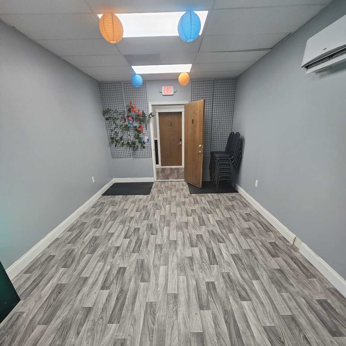 A narrow hallway with light gray wood-like flooring, a door at the end, and colorful lanterns hanging from the ceiling.