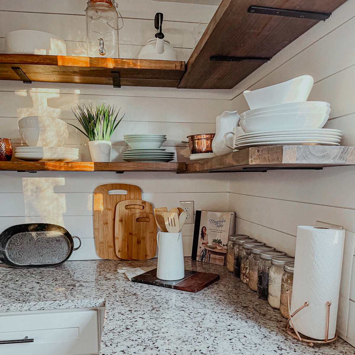 The kitchen features a granite countertop with wooden shelves above, displaying various dishware, utensils, and jars, along with a cutting board and a paper towel holder.