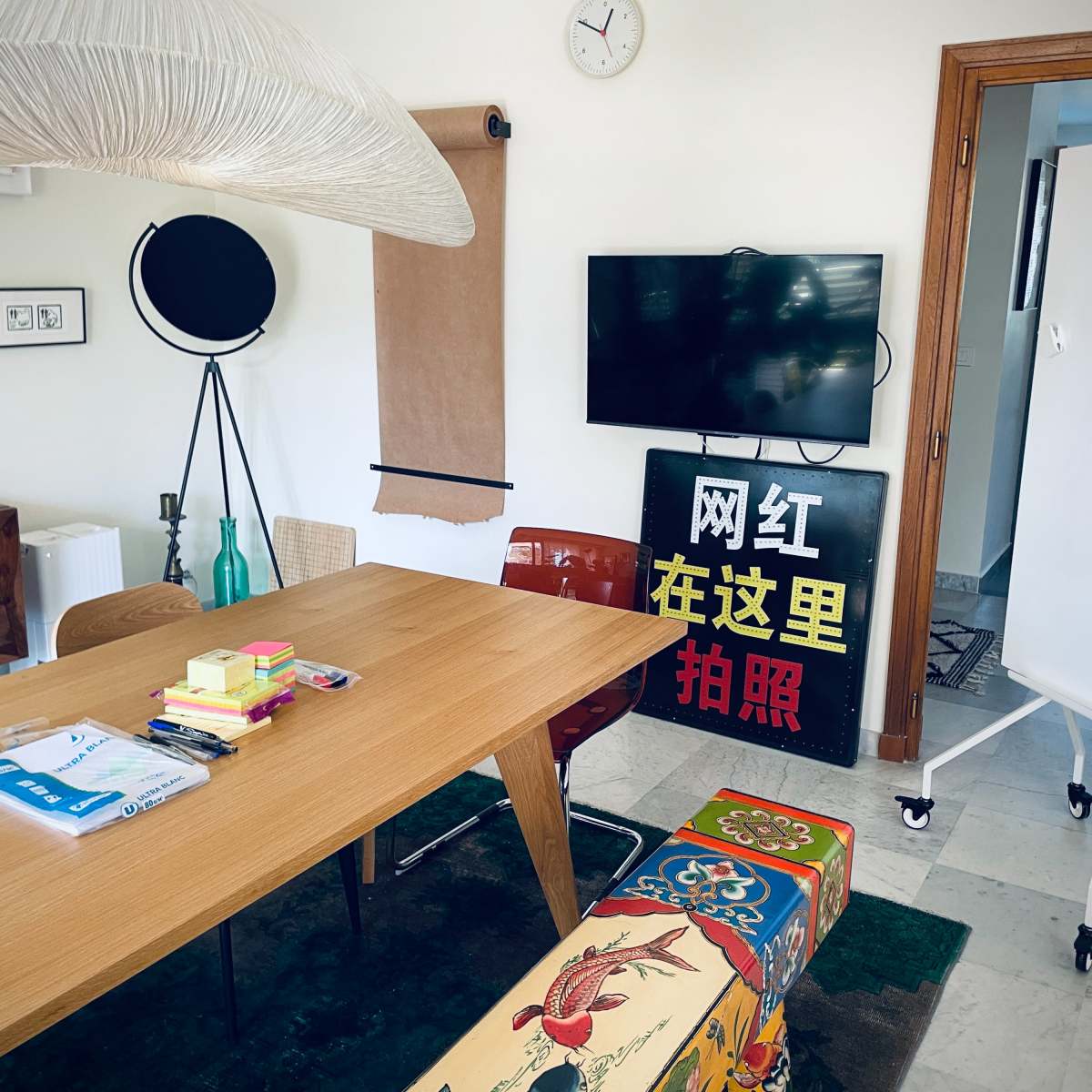 A bright meeting room features a wooden table surrounded by chairs, a large television, and a whiteboard, with decorative items and artwork on the walls.