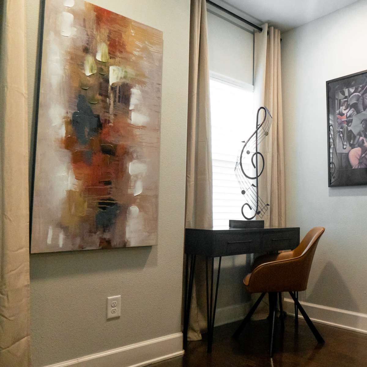 A small nook features a desk with a chair, flanked by two paintings on the walls, and a window with light-colored curtains.