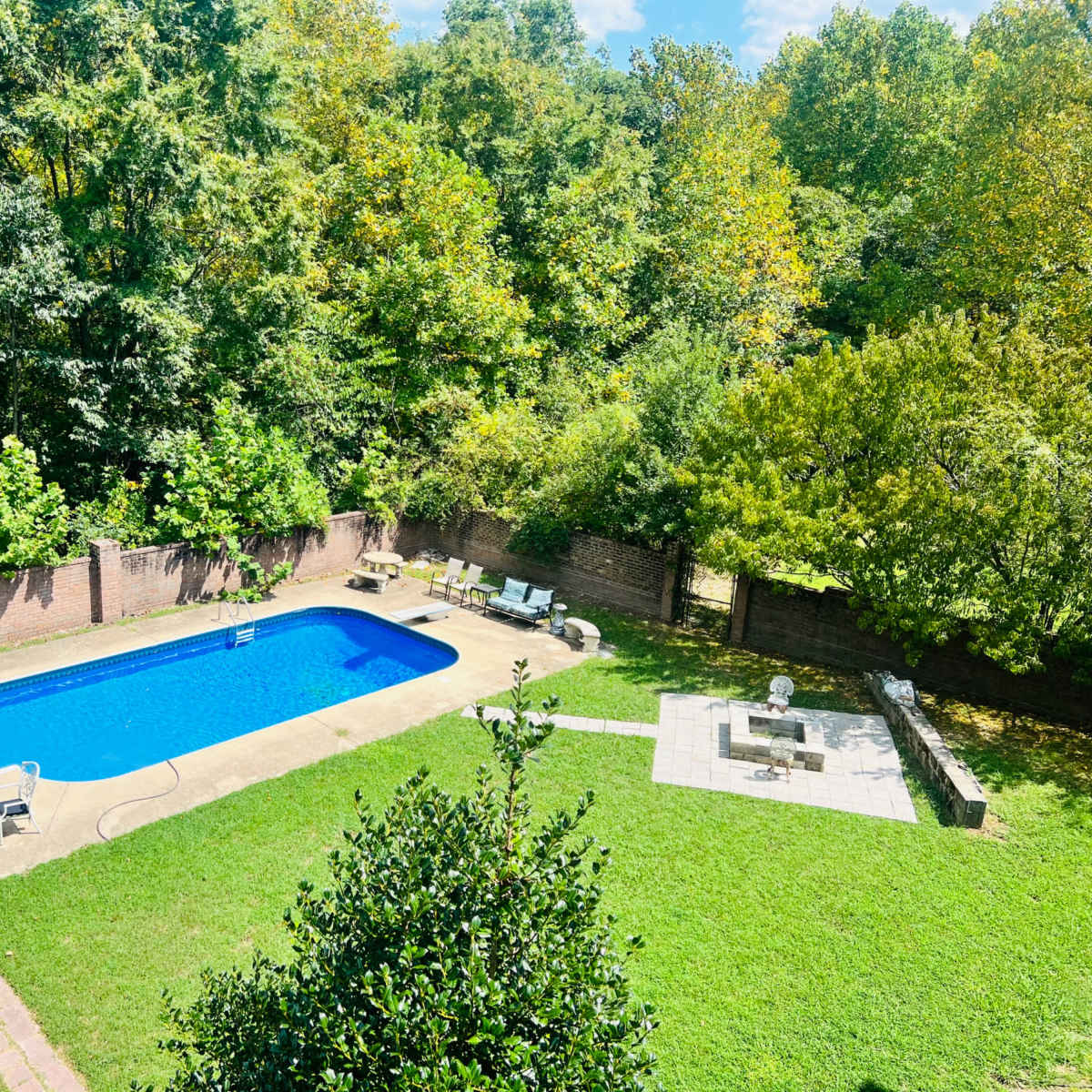 A rectangular swimming pool surrounded by a grassy area, patio furniture, and a stone fire pit, all enclosed by a brick wall and lush greenery.