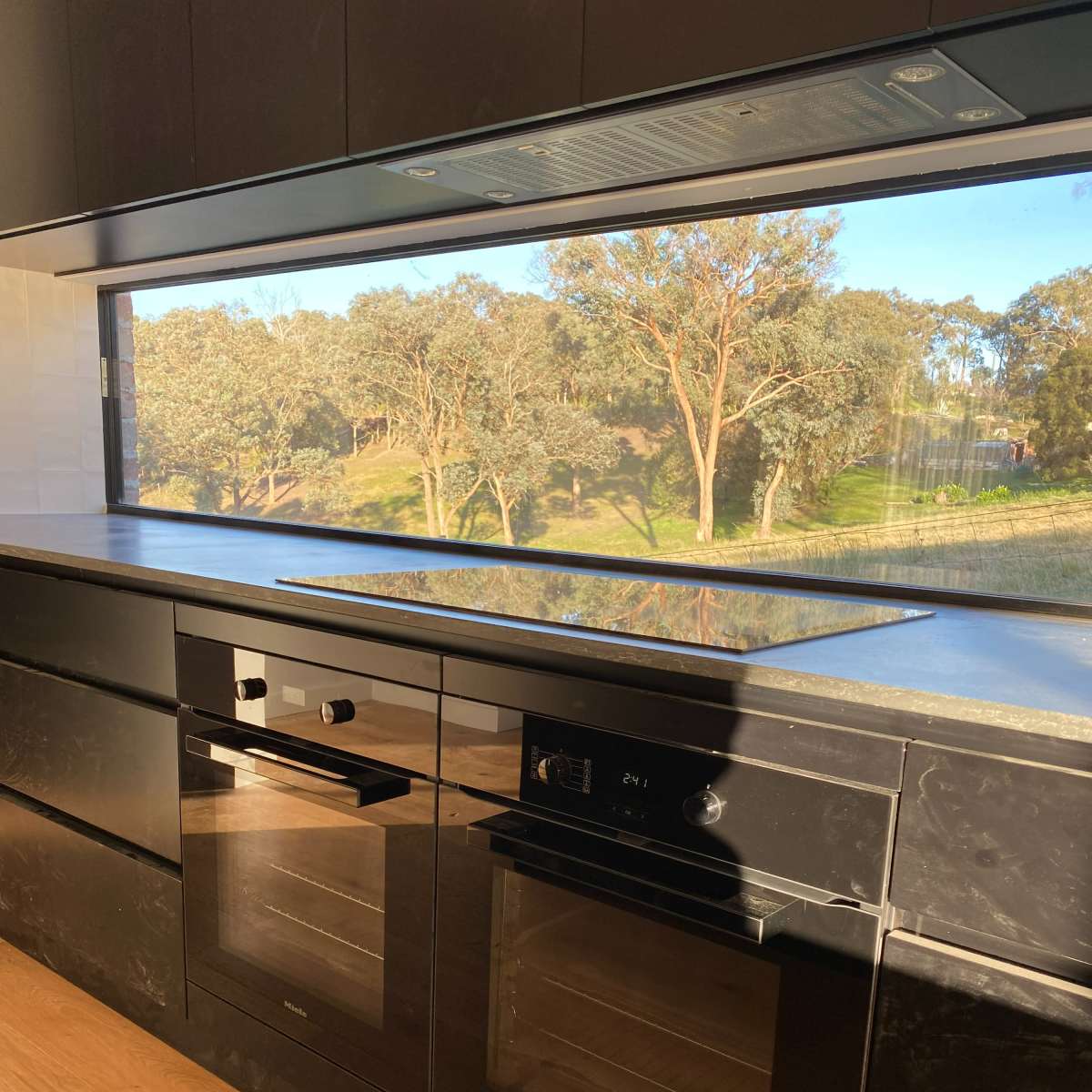 A modern kitchen with black cabinetry, built-in ovens, and a large window overlooking trees and greenery outside.