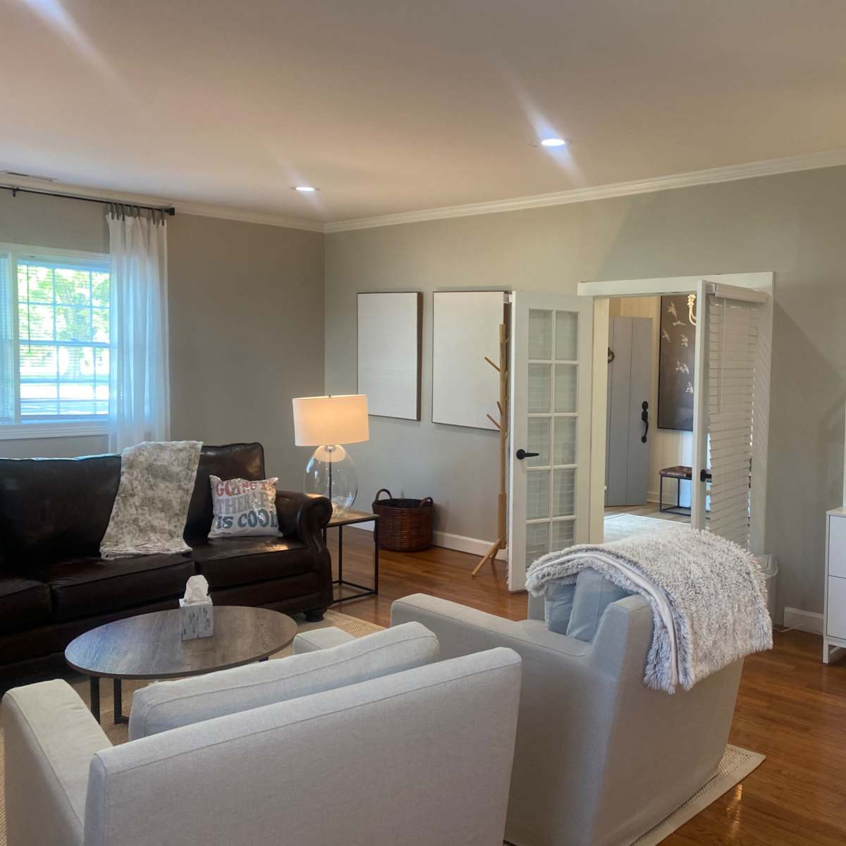 The room features a cozy living space with a dark sofa, light-colored armchairs, a white bookshelf, and sliding doors leading to another area.