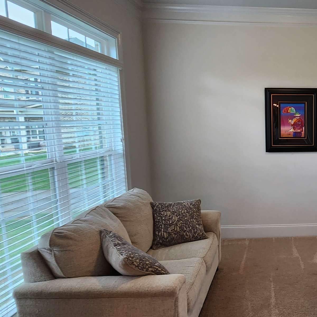 A small, well-lit room containing a beige sofa, a gray armchair, a side table, and two framed artworks on the wall.