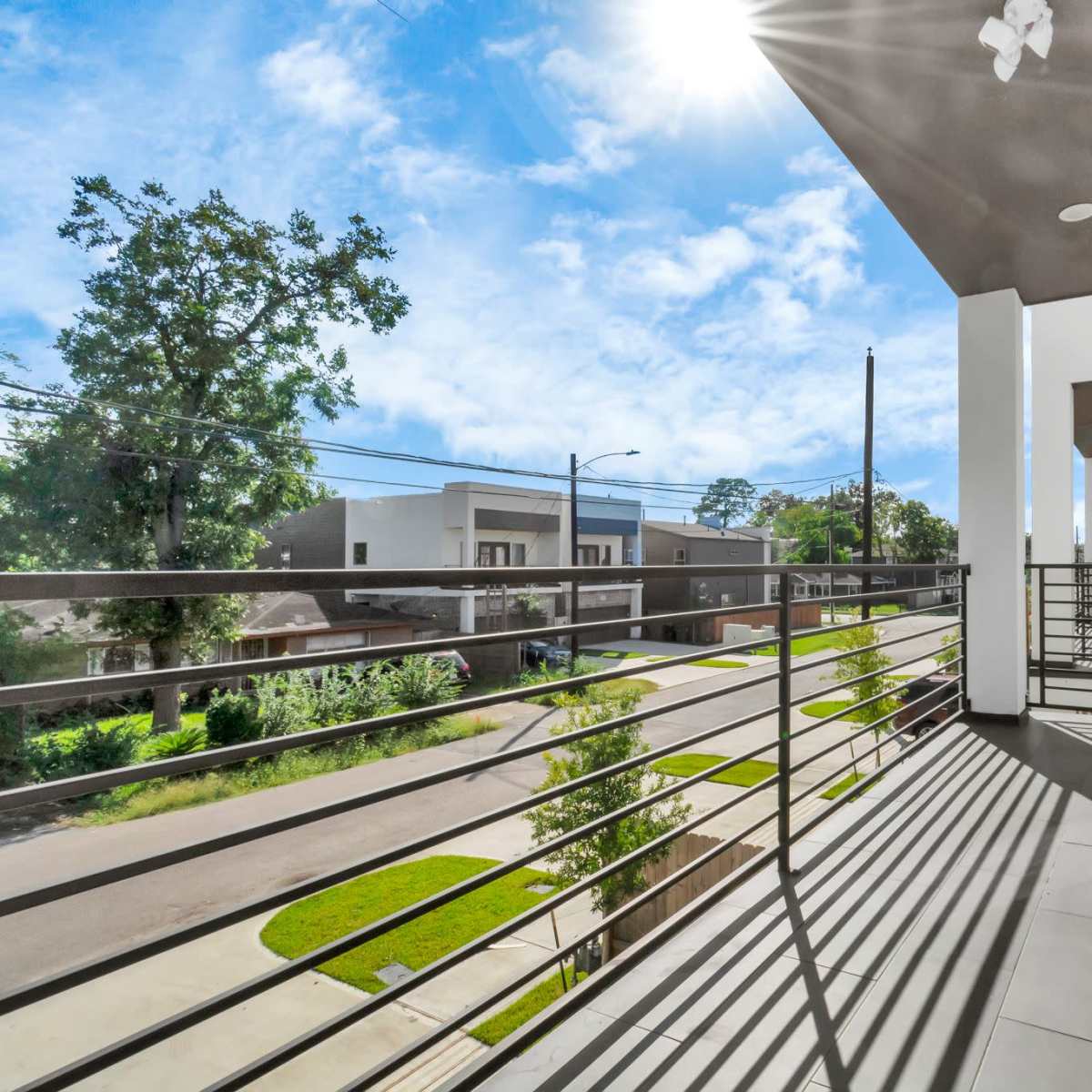 A balcony with a railing overlooking a street lined with trees and modern buildings under a bright sky.