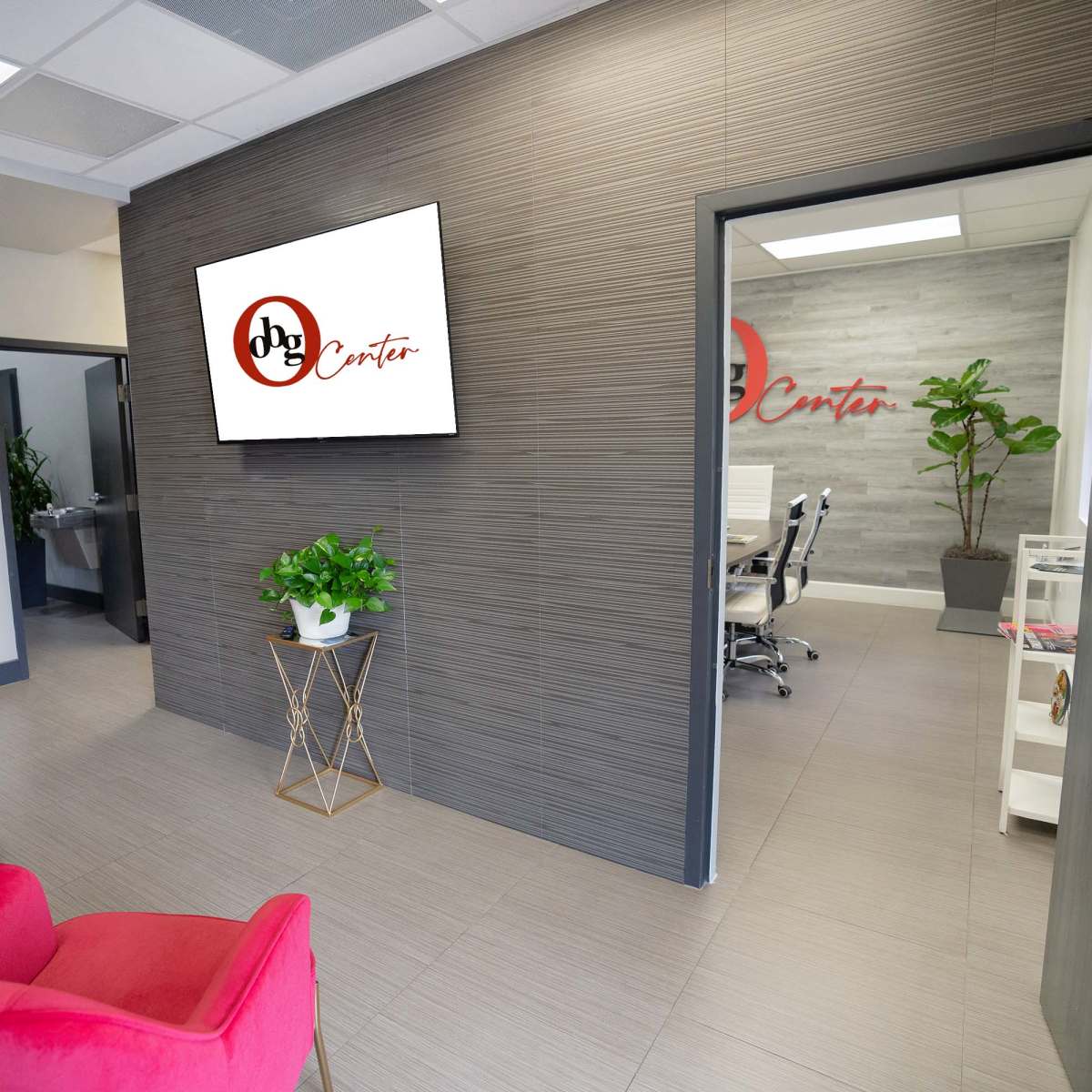 A modern reception area with a potted plant, a red chair, and a wall-mounted television displaying the logo of a center, leading to a conference room.