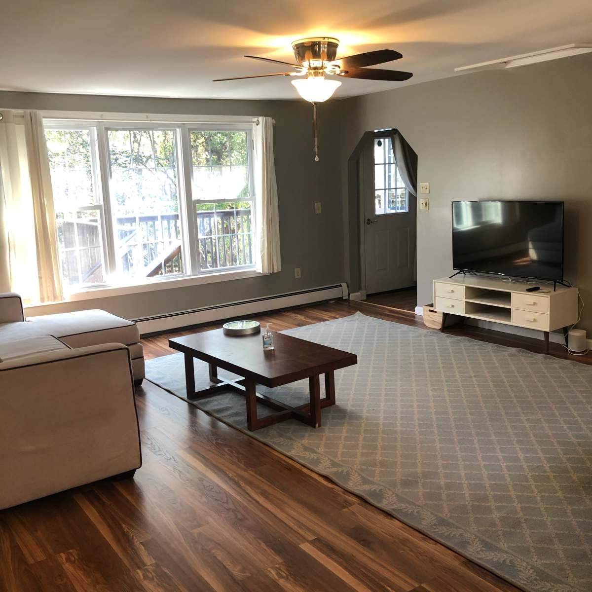 The living room features a beige sectional sofa, a wooden coffee table, a television on a white stand, and large windows that offer natural light.