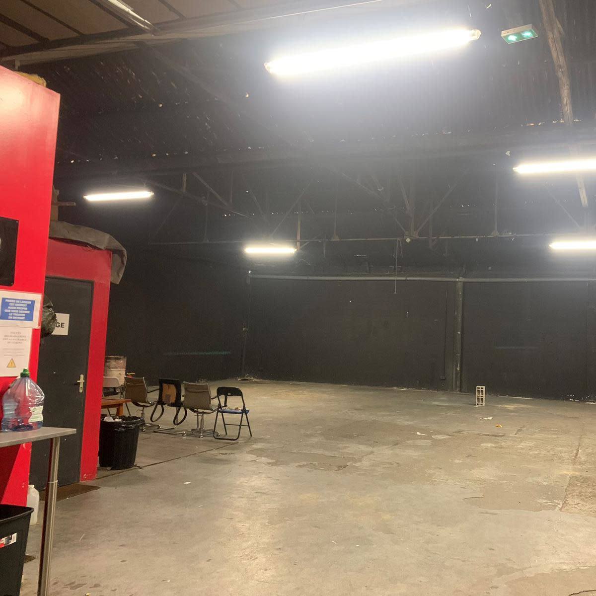 A spacious, dimly lit warehouse interior features black walls, a concrete floor, and various furniture items along the perimeter.