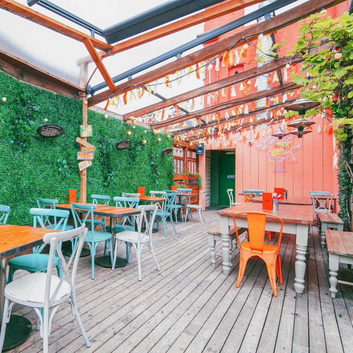A brightly lit outdoor dining area with wooden tables and colorful chairs, surrounded by greenery and hanging decorations.