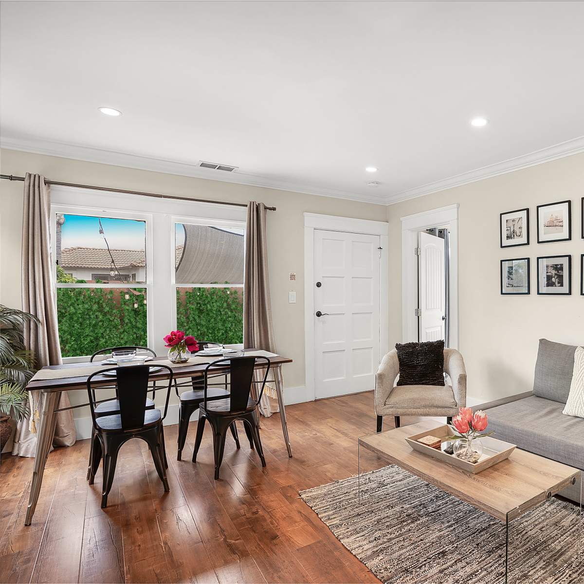 The living room features a gray sectional sofa, a wooden coffee table with flowers, framed pictures on the wall, and a dining area with a table and chairs by a window.